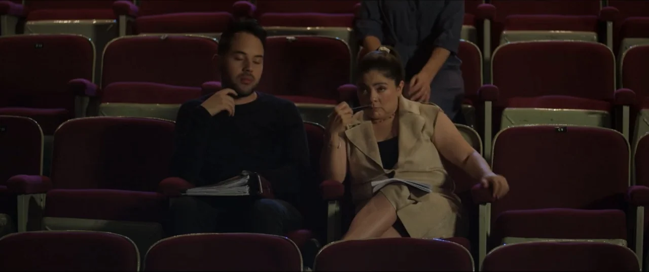 Can't Quit Mom (2022) movie still: medium — A rows of velvet theater seats fill a dark auditorium during a rehearsal. A man and a woman sit side by…