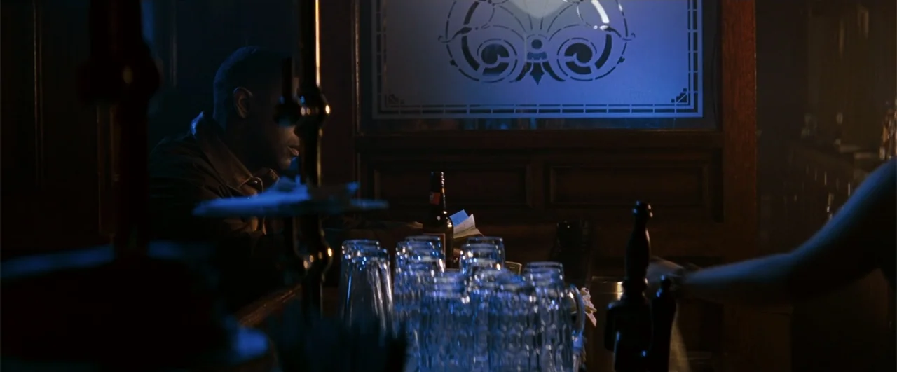 Fallen (2004) movie still: medium — A dark bar interior features a wooden counter and a glowing etched glass panel. A man sits in profile at the bar…