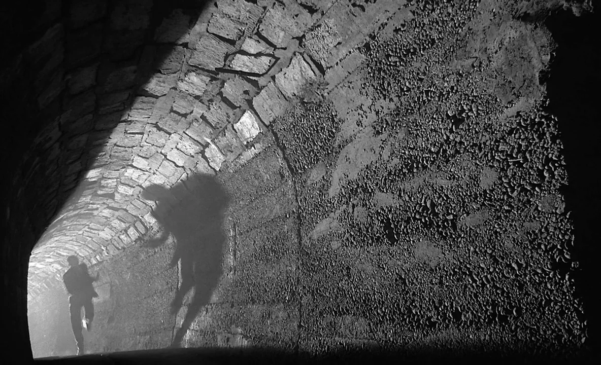 The Trial (1962) movie still: wide — A long curved tunnel constructed of uneven stone blocks and textured walls. A lone figure runs away from the…