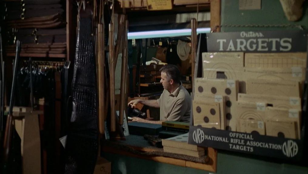 Targets (1968) movie still: medium — A man works behind a wooden counter in a store filled with supplies. Paper targets are displayed prominently on…