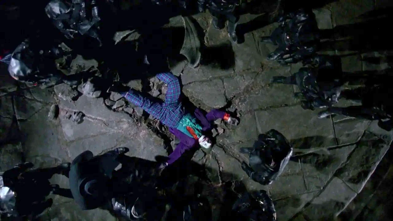 Batman (1989) movie still: wide — A stone-paved plaza at night is surrounded by dark figures in tactical gear. A man in purple and green suit lies…