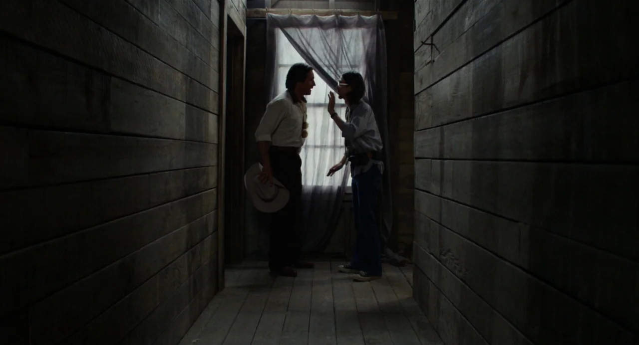 X (2022) movie still: fullbody — A narrow corridor features dark wood paneling on both walls and floorboards. Two individuals stand at the end of…