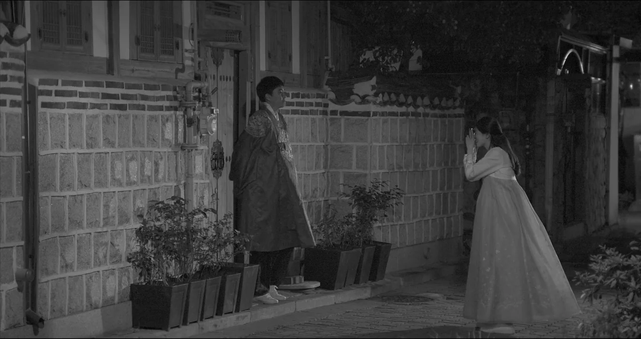 Grass (2018) movie still: fullbody — A stone-walled exterior of a Korean building features a wooden gate and potted plants. A woman photographs a…