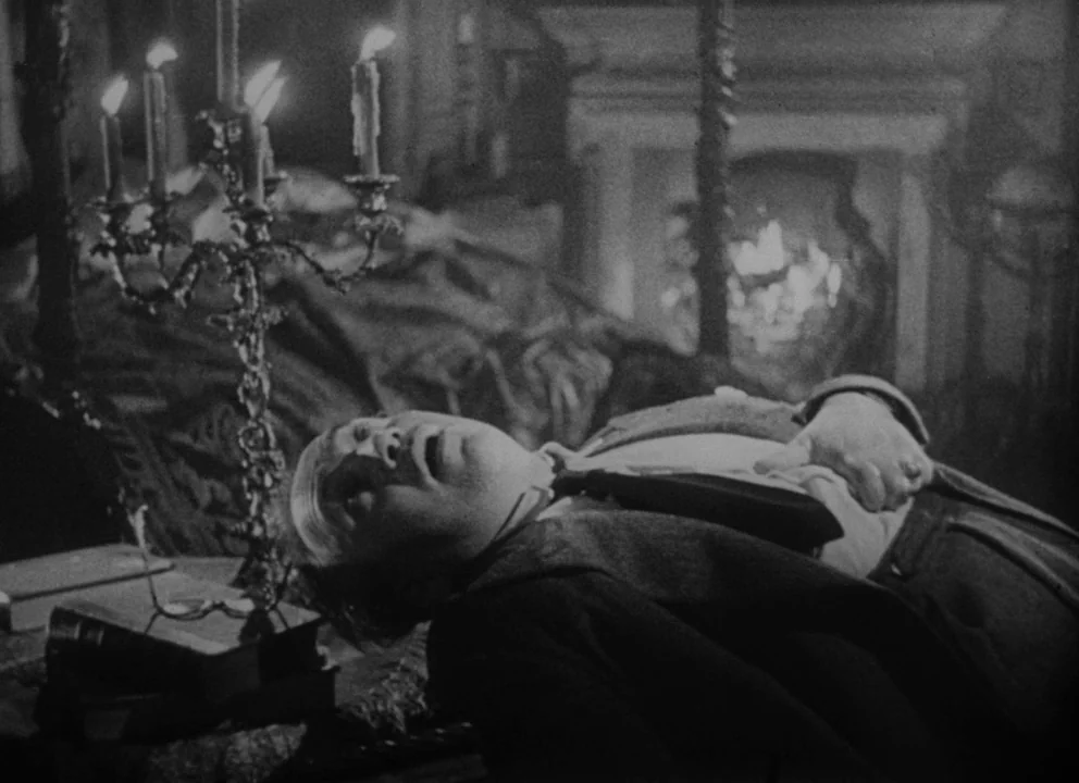 Dead of Night (1945) movie still: medium — A dimly lit classic study features a fireplace and a heavy wooden desk. An older man in a suit lies…