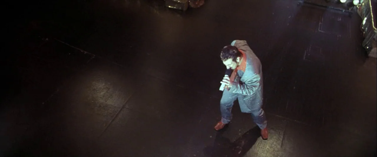 The Cooler (2003) movie still: wide — A dark wooden performance space with a single person standing in the center. The man performs while holding a…
