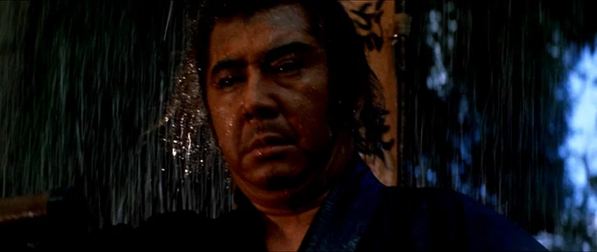 Lone Wolf and Cub: Sword of Vengeance (1972) movie still: closeup — A man stands outdoors under heavy rainfall during the night. Rain streaks across…