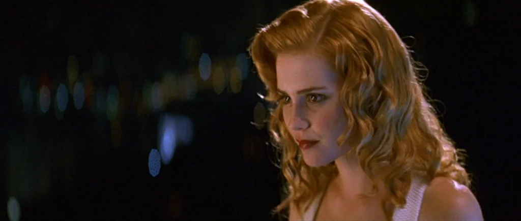 Where The Truth Lies (2005) movie still: closeup — A woman stands in darkness against a blurry night city view. She looks ahead while lit by a soft…