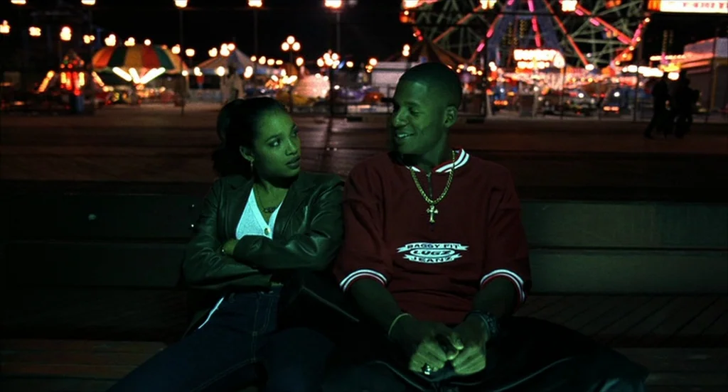 He Got Game (1998) movie still: medium — A night view of a boardwalk with colorful amusement park lights in the distance. Two young adults sit on a…