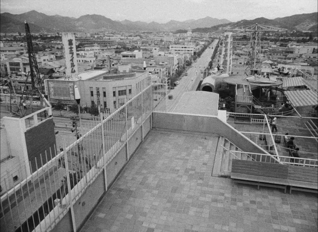 Hiroshima Mon Amour (1959) movie still: establishing — A wide view from a tiled rooftop overlooking a Japanese town with mountains beyond. The…