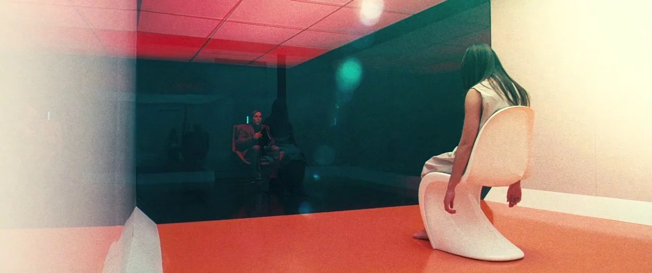 Beyond The Black Rainbow (2010) movie still: wide — A minimalist room with a bright orange floor and a large dark glass panel. A woman in a light…
