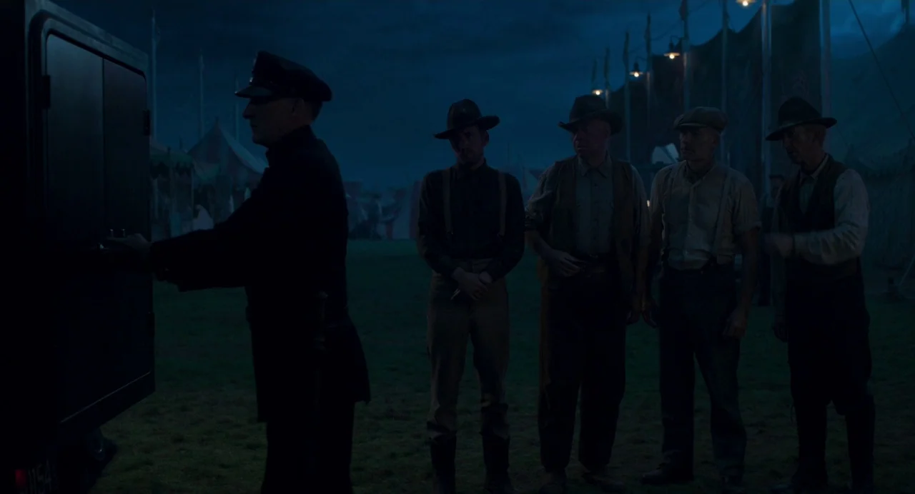 Dumbo (2019) movie still: fullbody — A dark night at a carnival grounds with large tents looming in the back. A police officer opens the back of a…