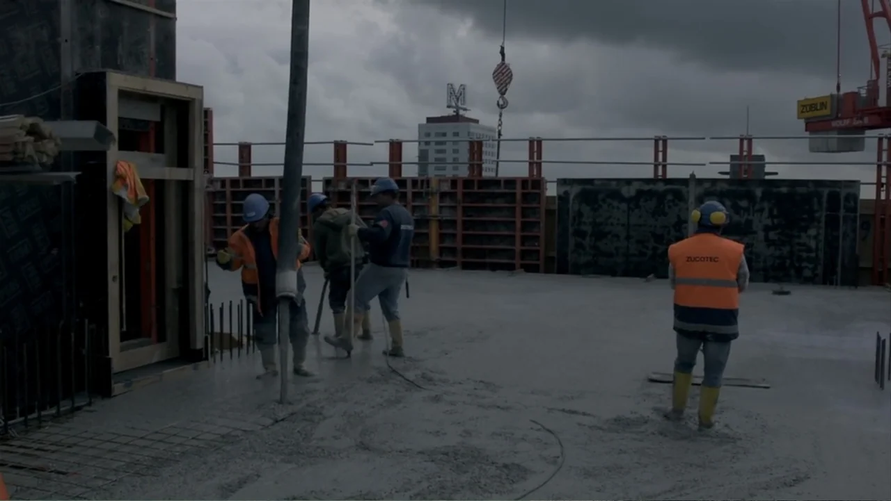 REM (2019) movie still: wide — An outdoor construction site on a high-rise floor under a cloudy grey sky. Several workers in high-visibility gear…
