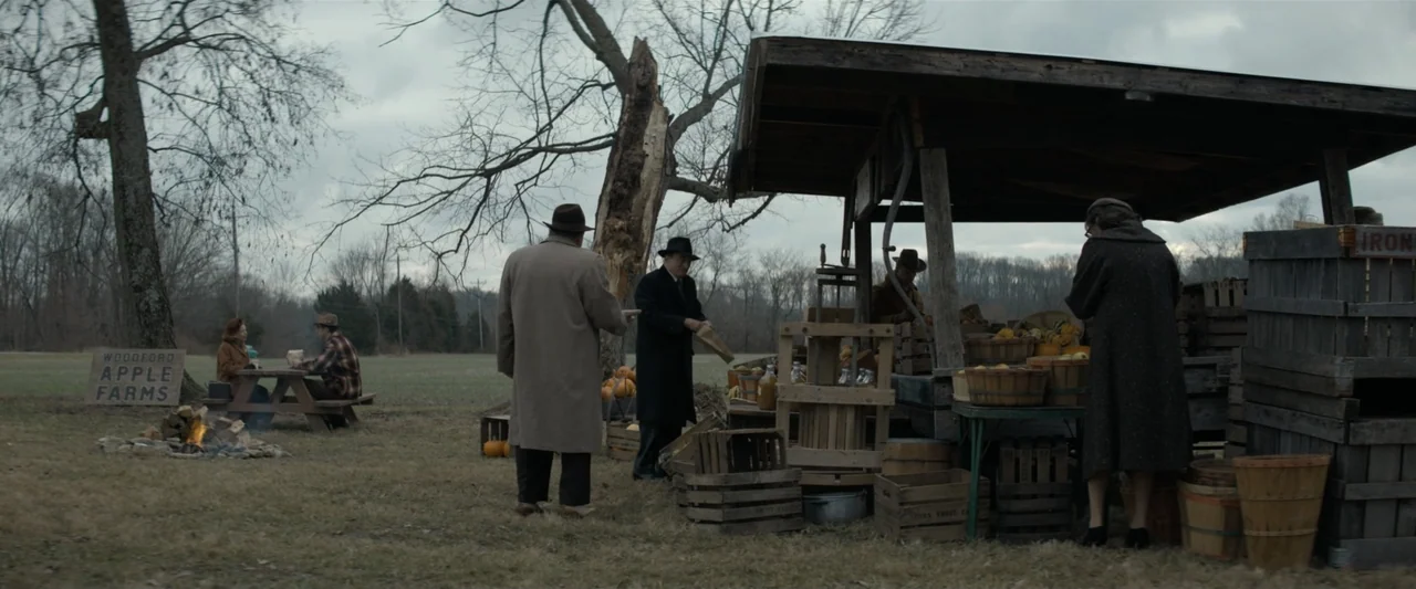 The Alto Knights (2025) movie still: wide — A rural apple farm features a weathered wooden produce stand under an overcast sky. Several individuals…