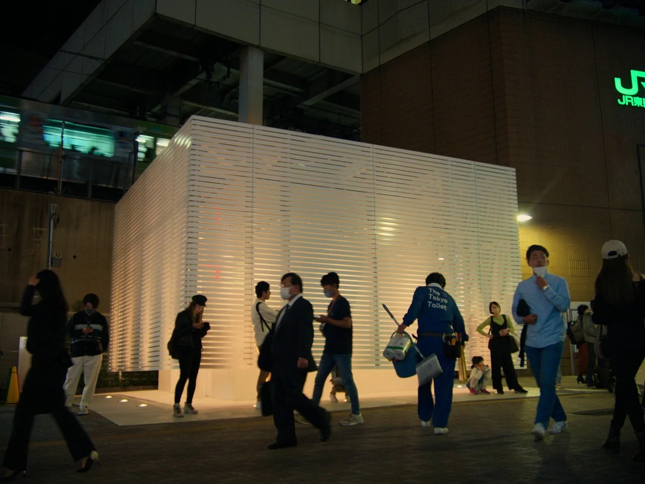 Perfect Days (2023) movie still: wide — An urban plaza at night features a modern white structure made of thin horizontal slats. Pedestrians walk…