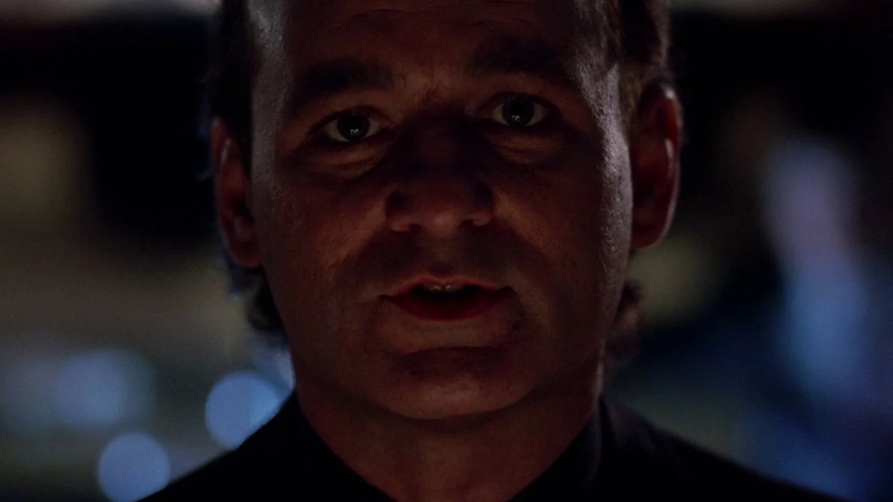 Scrooged (1988) movie still: closeup — A dimly lit interior space with dark shadows concealing the surroundings. A man faces the camera directly…