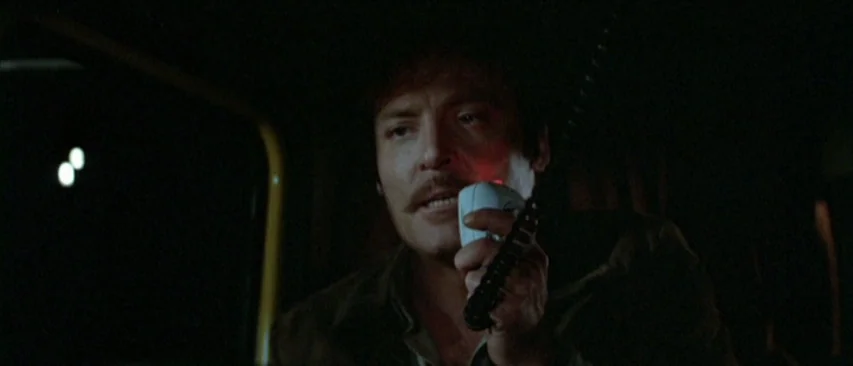 Roadgames (1981) movie still: closeup — A man sits inside a dimly lit vehicle cabin during the nighttime. He holds a CB radio microphone close to…