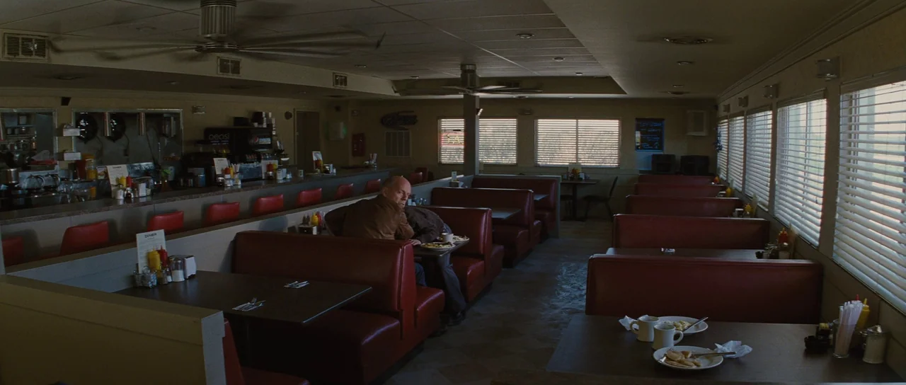 Looper (2012) movie still: wide — A dimly lit classic diner interior features several rows of red vinyl booths. A lone man in a brown leather jacket…