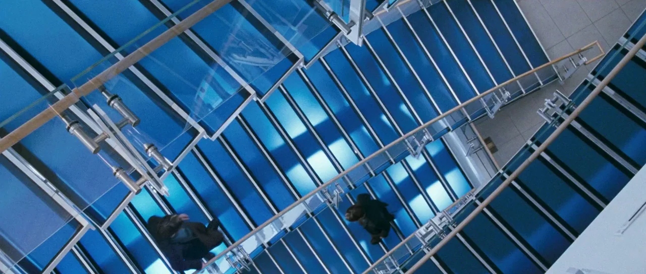 Stay (2005) movie still: wide — A modern building interior features a complex, multi-level glass staircase with backlit blue panels. Two people are…