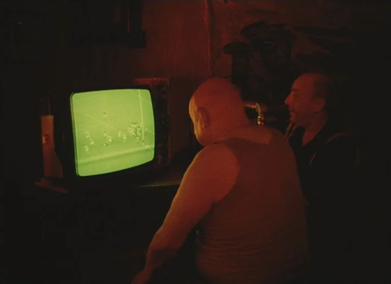 A Visitor to a Museum (1989) movie still: medium — A dimly lit room with walls cast in deep red shadows from ambient darkness. Two older men sit…