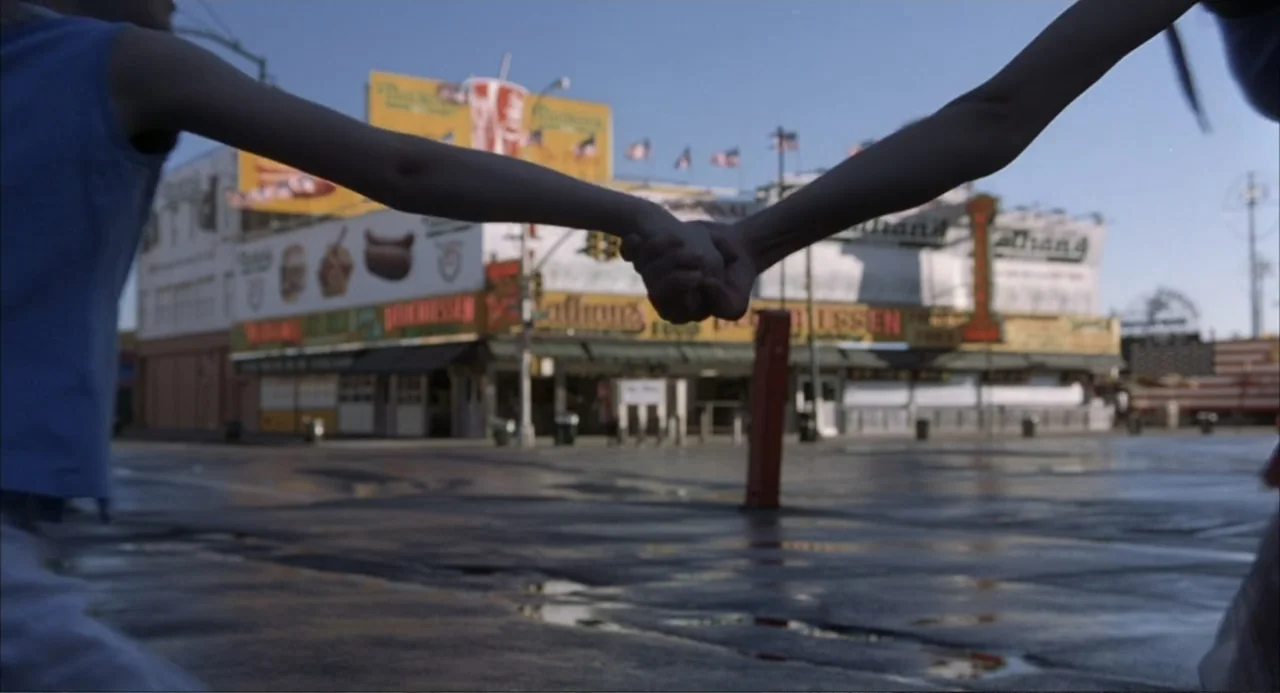 Uptown Girls (2003) movie still: medium — A paved boardwalk area displays large food signage and American flags in the distance. Two people reach…