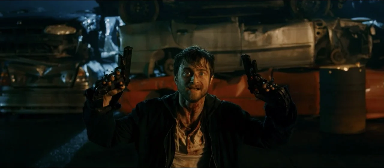 Guns Akimbo (2020) movie still: medium — A dark outdoor junkyard stacked with rusted automobiles at night. A disheveled man kneels holding guns…