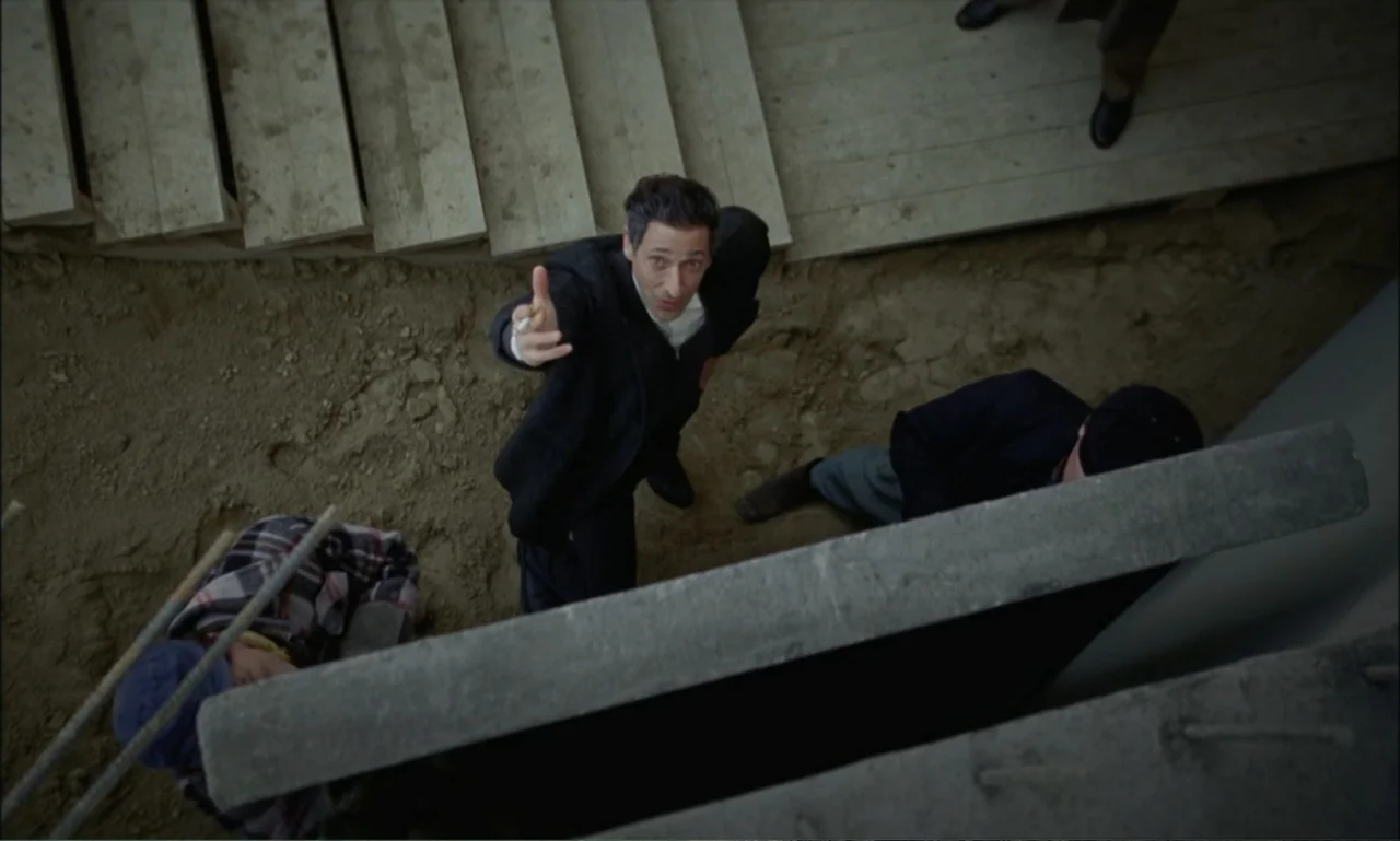 The Brutalist (2024) movie still: wide — A dirt-covered construction site with concrete foundations and wooden steps visible from above. A man in a…