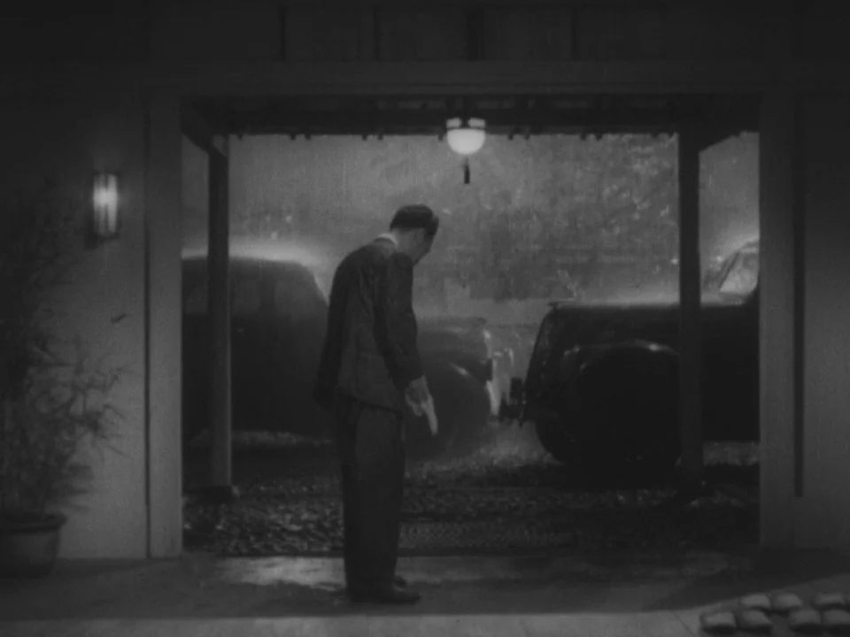 Stray Dog (1949) movie still: fullbody — A man stands in a dark residential carport during a heavy night rainstorm. Two vintage automobiles are…