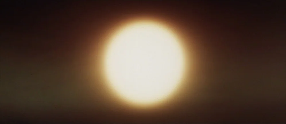 Dune (1984) movie still: establishing — A clear, expansive sky displays a bright sun at the center of the frame. The horizon is shrouded in a dark,…