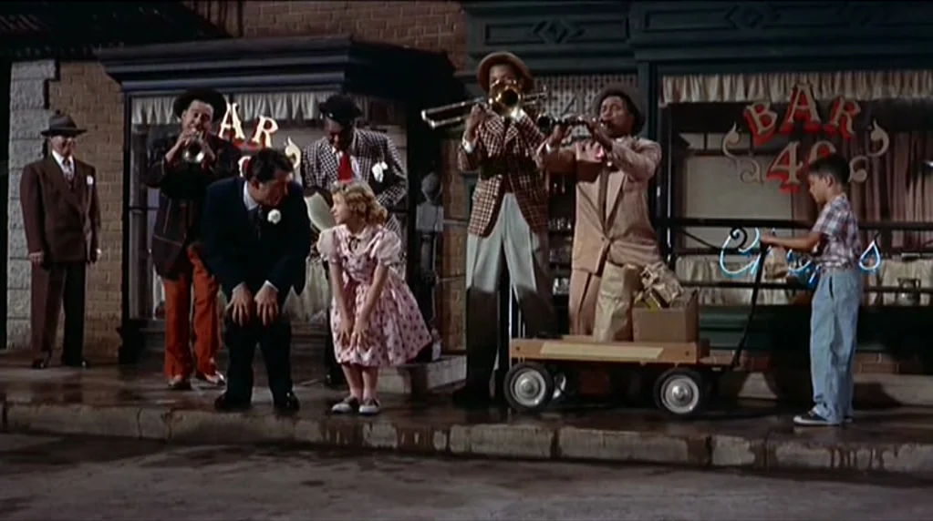 Artists and Models (1955) movie still: wide — A sidewalk in front of a brick building featuring shops with glass storefronts. A group of men in…