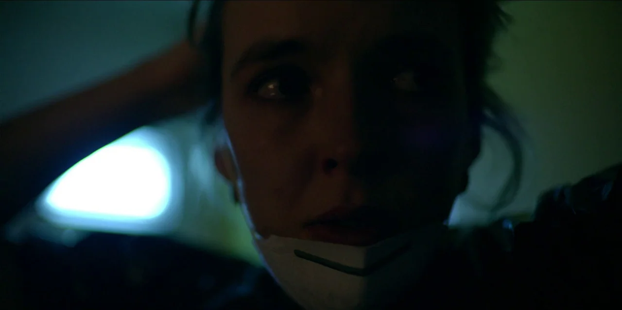 Help (2021) movie still: closeup — A dimly lit room with cool shadows obscuring the background. A woman looks off-camera while wearing a white face…