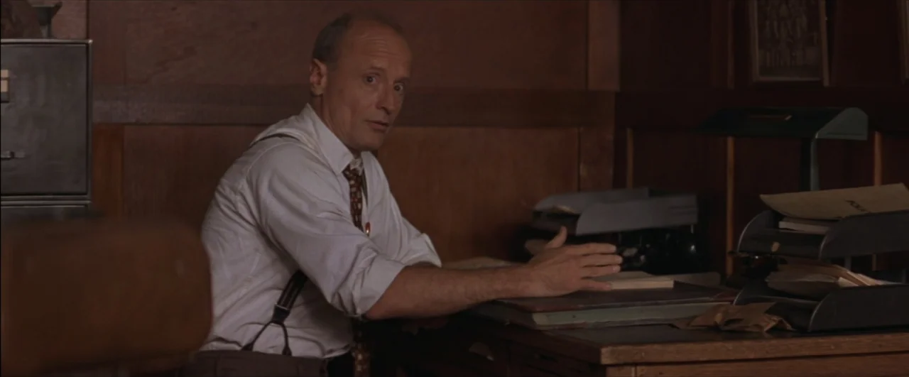 This Boy's Life (1993) movie still: medium — A 1940s wood-paneled office contains a desk with paper trays and documents. A balding middle-aged man…