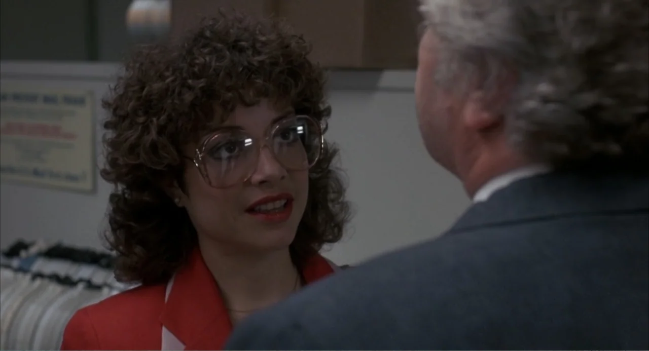 F/X (1986) movie still: closeup — A brightly lit retail environment features racks of clothing in the background. A woman in a red blazer engages in…