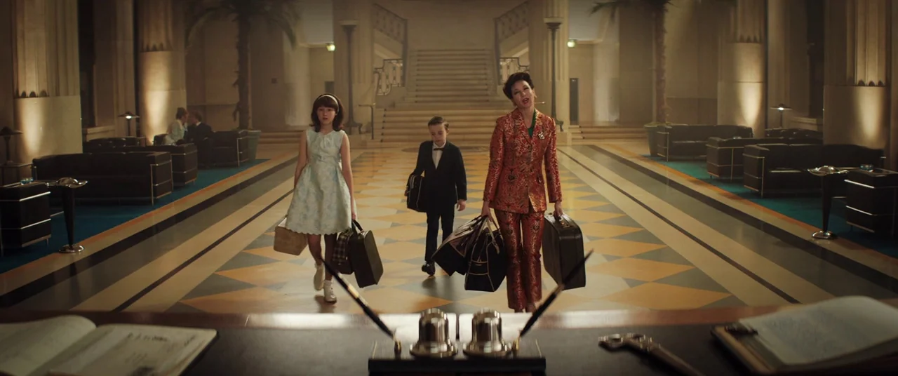 Judy (2019) movie still: fullbody — A spacious, opulent lobby featuring checkered tile floors and grand staircases. A woman and two children walk…
