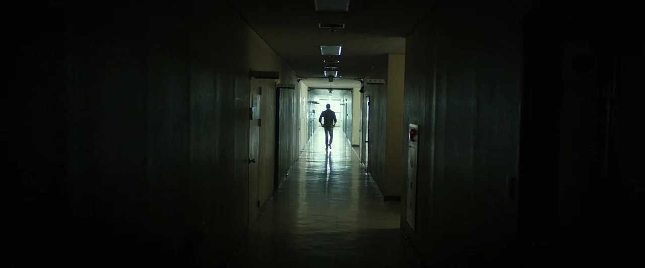 Believer (2018) movie still: wide — A long, dimly lit building corridor with multiple closed doors along beige walls. A lone man walks away from the…