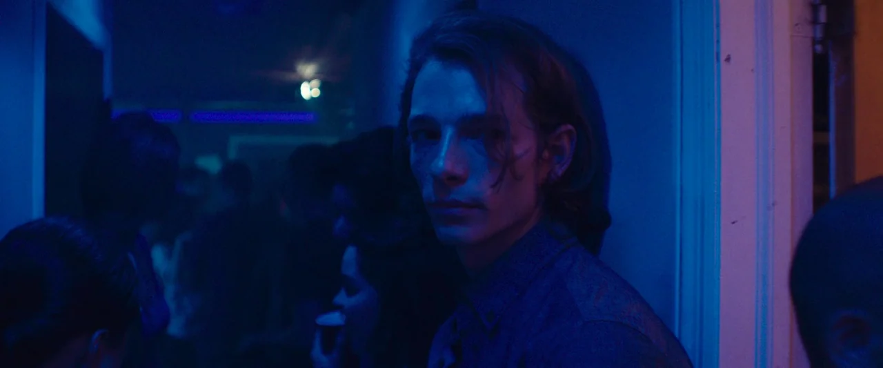 Wildling (2018) movie still: medium — A crowded, dimly lit nightclub interior with blue neon light and blurred figures. A young man with long hair…