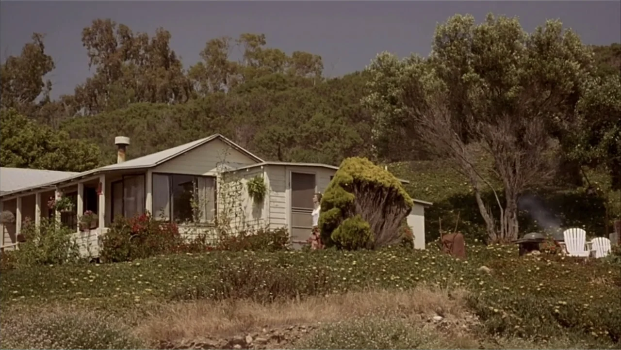 Ajuste de Cuentas (2016) movie still: wide — A small light-colored house sits on a grassy hill surrounded by trees. A woman stands by the entrance…