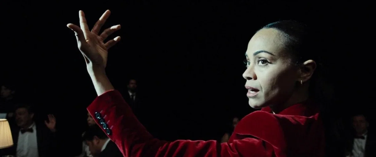 Emilia Pérez (2024) movie still: medium — A dark, dimly lit banquet hall or dining space at night. A woman in a red jacket gestures with her hand…