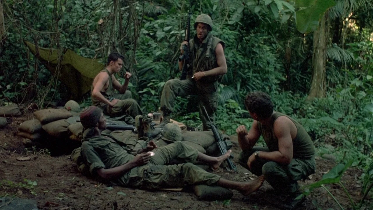 Platoon (1986) movie still: wide — A group of soldiers occupies a makeshift camp behind sandbag walls in a dense jungle. Four men in military gear…