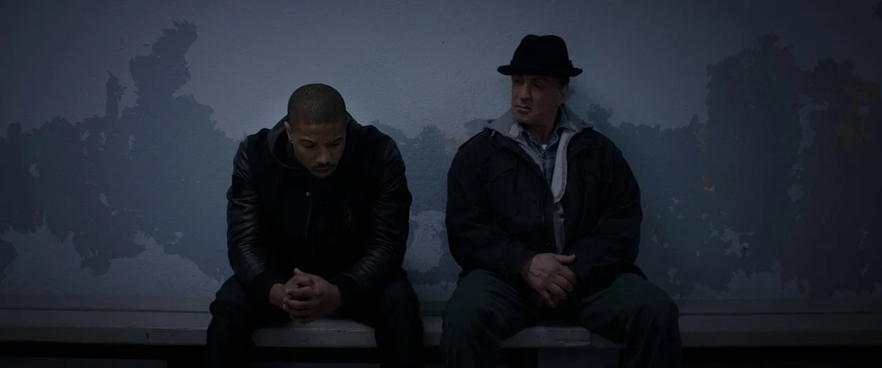 Creed (2015) movie still: medium — A long bench sits before a peeling, distressed blue-grey wall surface. Two men sit together looking forward in a…