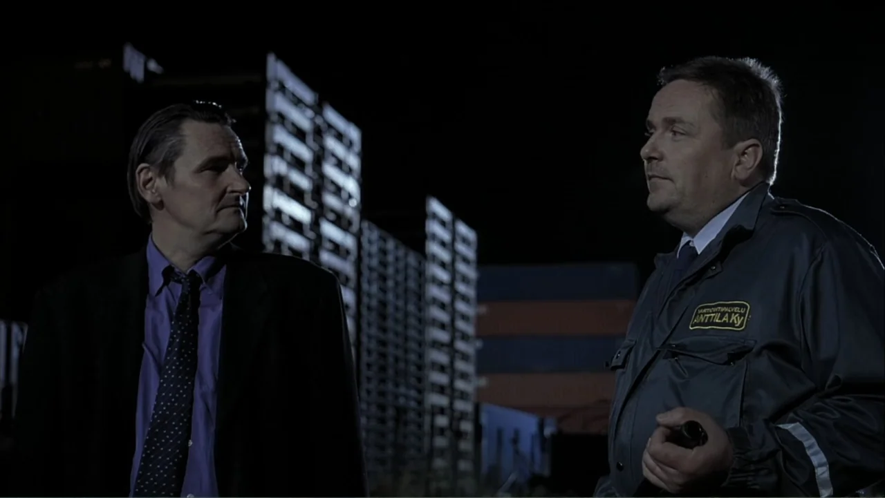 The Man Without a Past (2002) movie still: medium — A dark, nighttime industrial area features stacks of shipping containers in the background. Two…