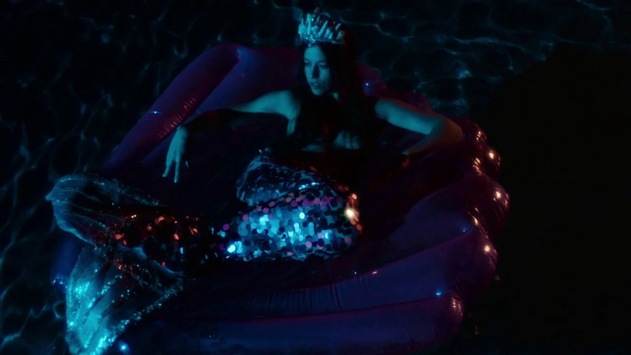 Snowbound (2017) movie still: medium — A woman wears a mermaid costume while lounging on a giant inflatable seashell. The figure is submerged in…