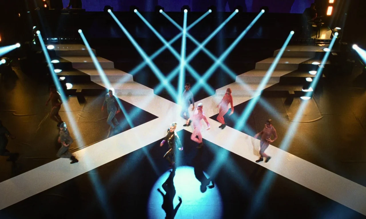 Vox Lux (2018) movie still: wide — A high-angle view of a dark stage featuring intersecting white pathways forming an X. Performers in colorful…