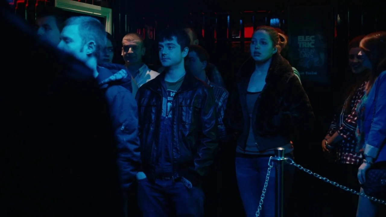 A Date for Mad Mary (2016) movie still: medium — A crowded, dimly lit indoor space with black tiled walls and neon lighting accents. A group of…
