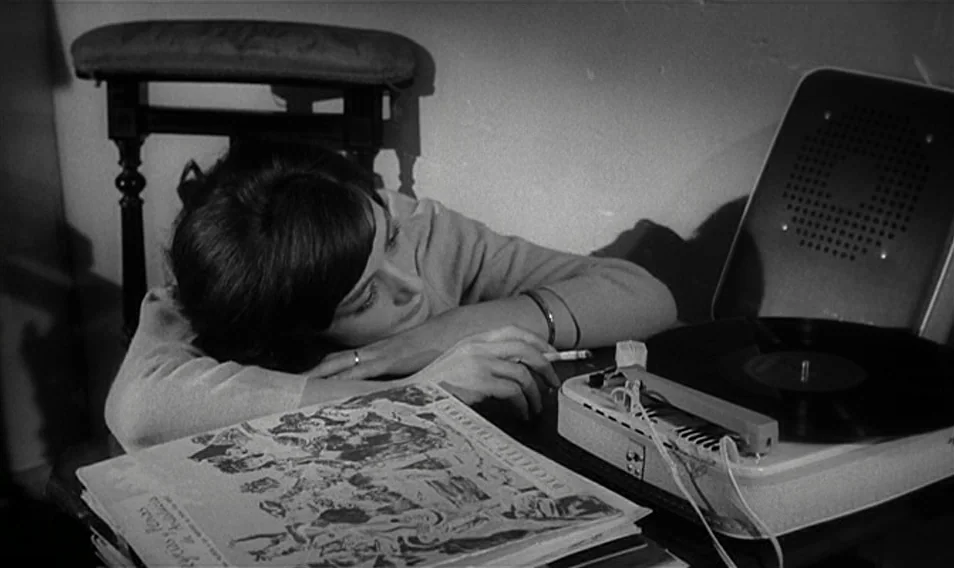Thérèse Desqueyroux (1962) movie still: medium — A dimly lit room interior features a young woman resting by a record player. She lies with her head…