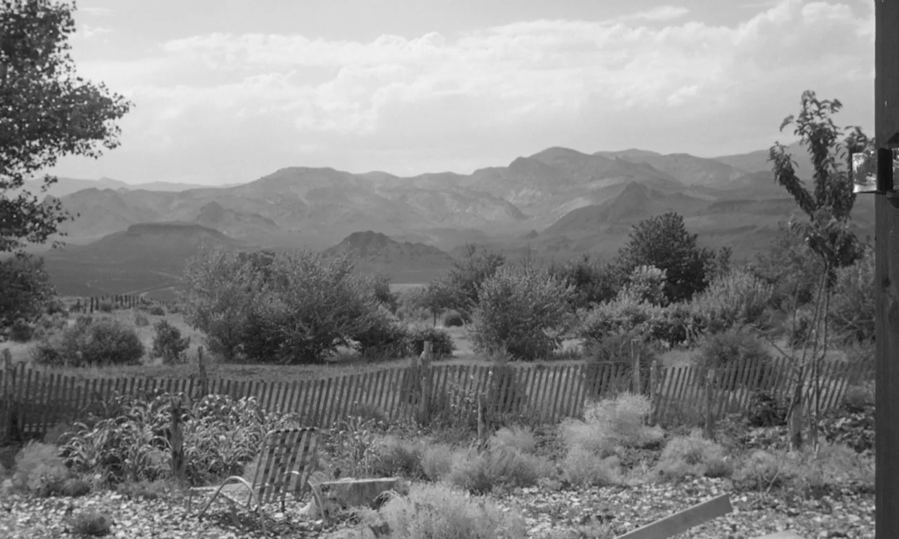 The Misfits (1961) movie still: establishing — A rural yard with a wooden picket fence overlooks vast, layered desert mountain ranges. A single…