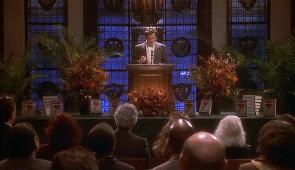 The Mirror Has Two Faces (1996) movie still: wide — A formal event takes place in an auditorium with dark wood paneling and windows. A man stands at…