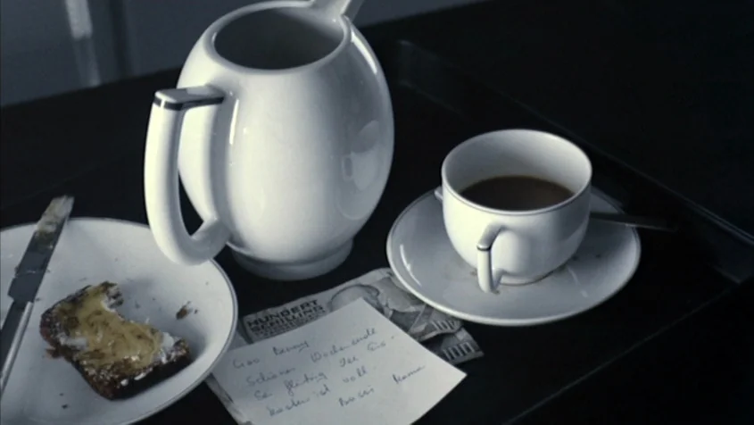 Benny’s Video (1992) movie still: closeup — A breakfast tray sits on a black surface holding white porcelain dinnerware. A handwritten note rests…
