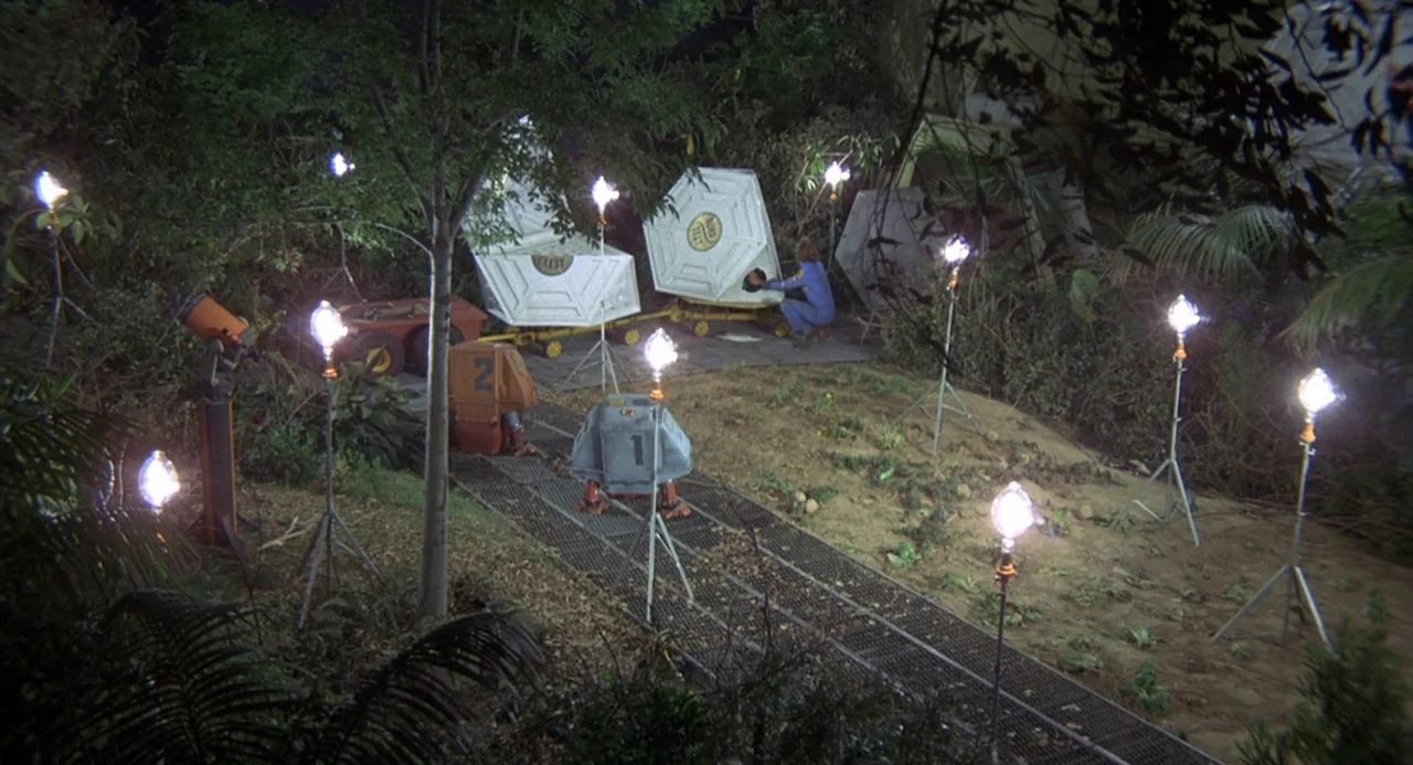Silent Running (1972) movie still: wide — A dark outdoor space features metallic containers and scattered portable lamps on thin metal stands. A…
