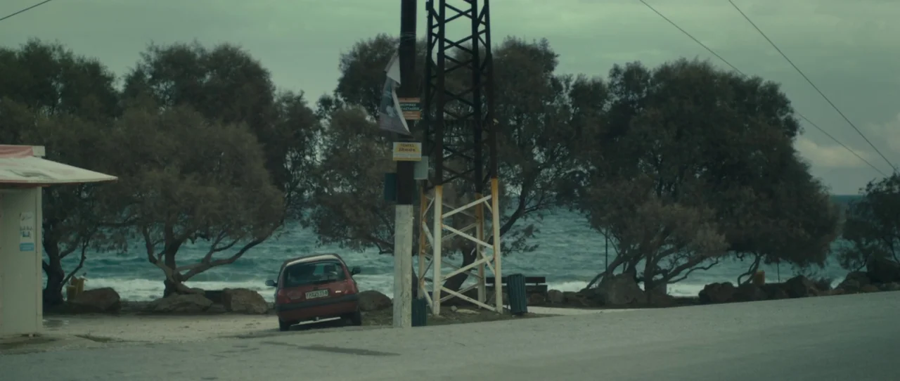 Alps (2011) movie still: wide — A coastal road overlook features a small hatchback parked beside a large metal pylon. Wispy trees and a turbulent…