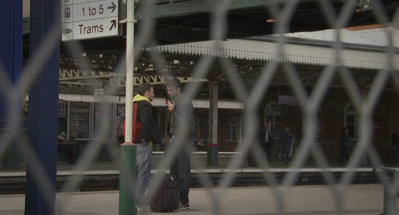 Weekend (2011) movie still: fullbody — An outdoor train station platform with metal fencing in the foreground. Two men stand facing each other while…
